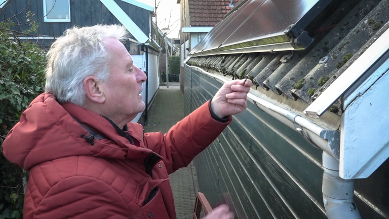 Een oudere man in een rode jas controleert een dakgoot met zonnepanelen erboven, in een smalle steeg.