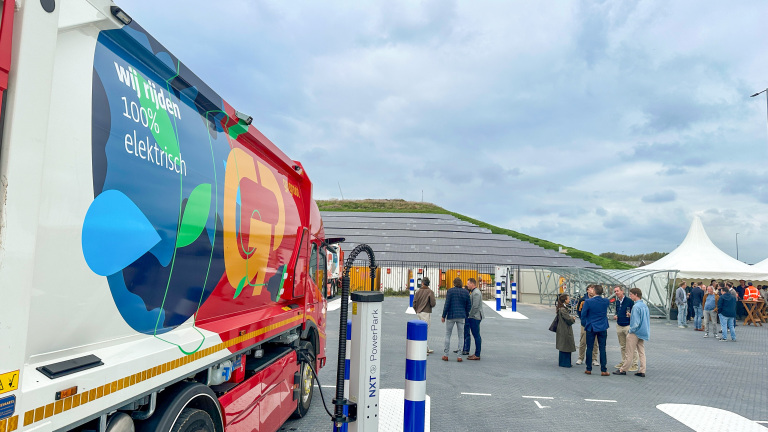 Vrachtwagen met tekst “Wij rijden 100% elektrisch” bij laadpalen, met groep mensen op parkeerterrein
