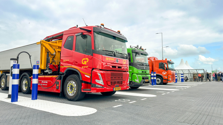 Drie vrachtwagens (rood, groen en oranje) geparkeerd bij een laadpunt op een parkeerterrein, met mensen op de achtergrond