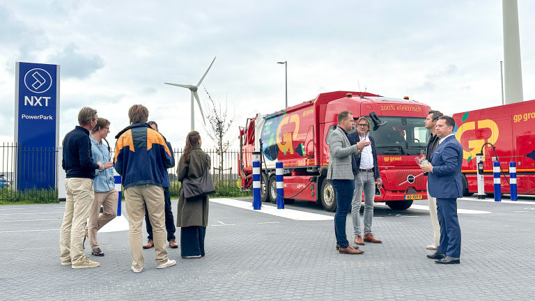 Groep mensen praat bij een laadpunt naast een rode vrachtwagen met tekst 100% elektrisch en een windturbine op de achtergrond