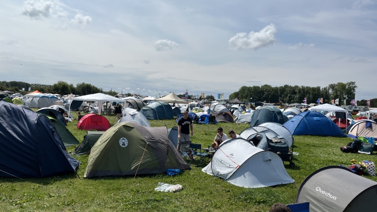 Veelzijdig kampeerterrein bij een festival, vol met kleurrijke tenten en enkele mensen die samen genieten, omgeven door een open veld en een heldere lucht.