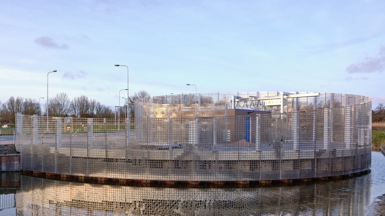 Een ronde structuur met een rastervormig metalen hekwerk, gelegen aan de rand van een waterpartij, onder een heldere hemel.