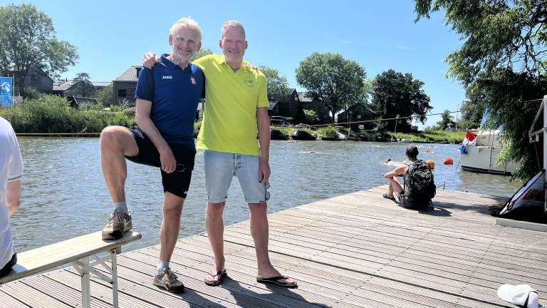 Twee mannen staan samen op een houten steiger aan het water op een zonnige dag, met zomerse kleding en omringd door groene bomen en enkele gebouwen op de achtergrond.
