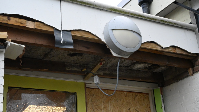 Vervallen gebouw met gebroken ramen en een dichtgetimmerde deur, omgeven door kapotte materialen op de grond.