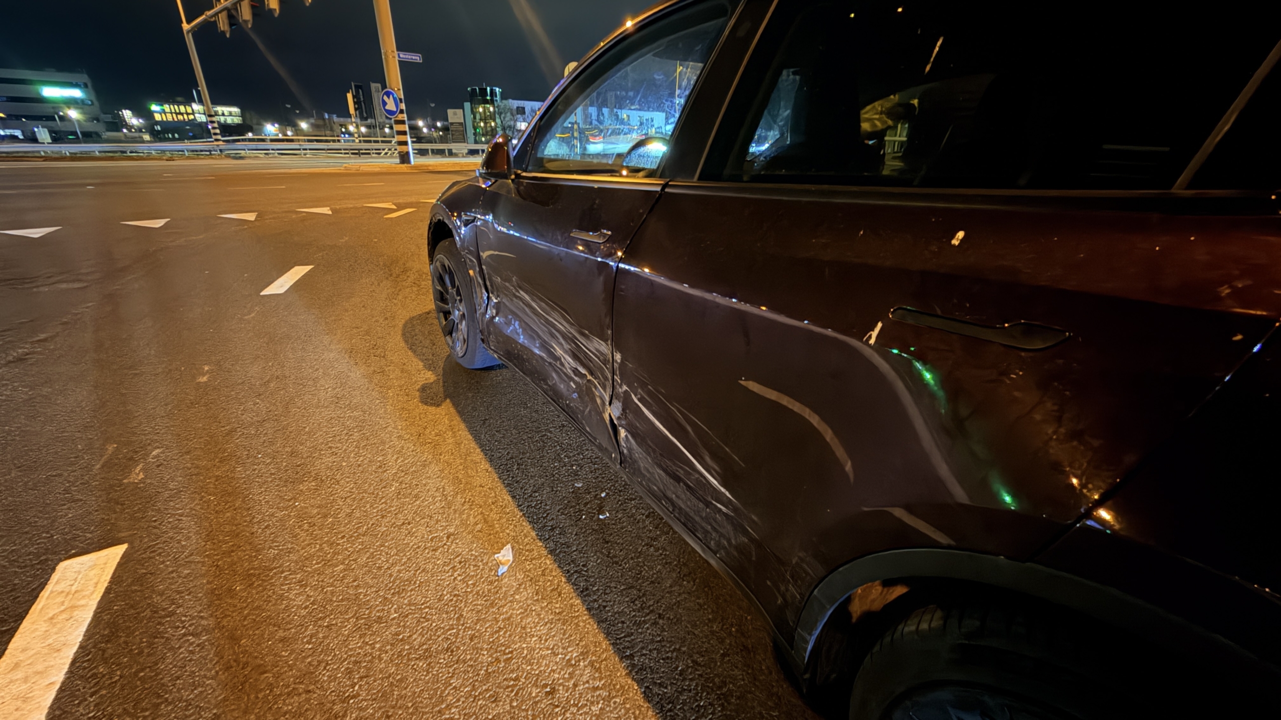 Auto met schade aan de voorkant op een weg 's nachts, met een politieagent en een verkeerskegel in de achtergrond.