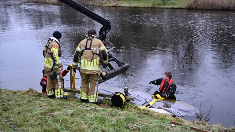 Brandweerlieden en een duiker halen een auto uit het water met behulp van een kraan langs een oever.
