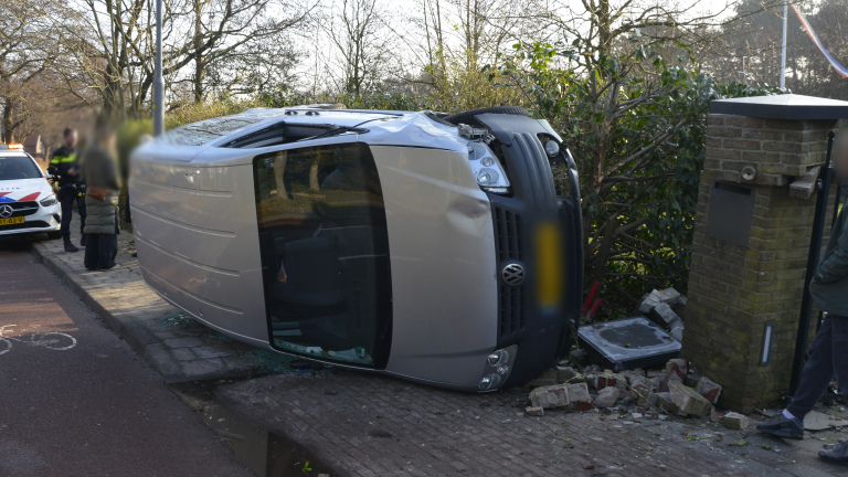 Auto ligt op zijn zijkant naast een kapotte muur; politieauto en mensen op de achtergrond.
