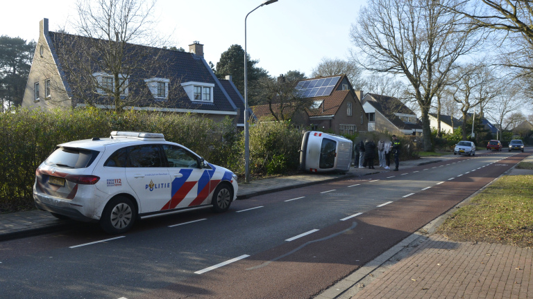 Auto ligt op zijn zijkant naast een kapotte muur; politieauto en mensen op de achtergrond.