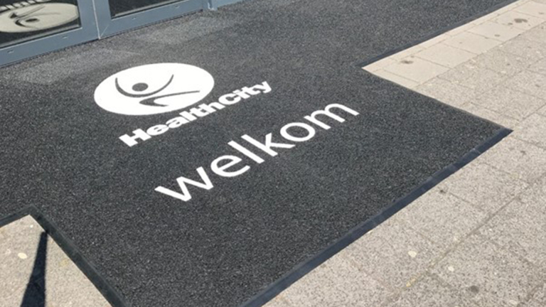Zwarte deurmat met een logo en tekst "HealthCity welkom" bij de ingang van een gebouw.