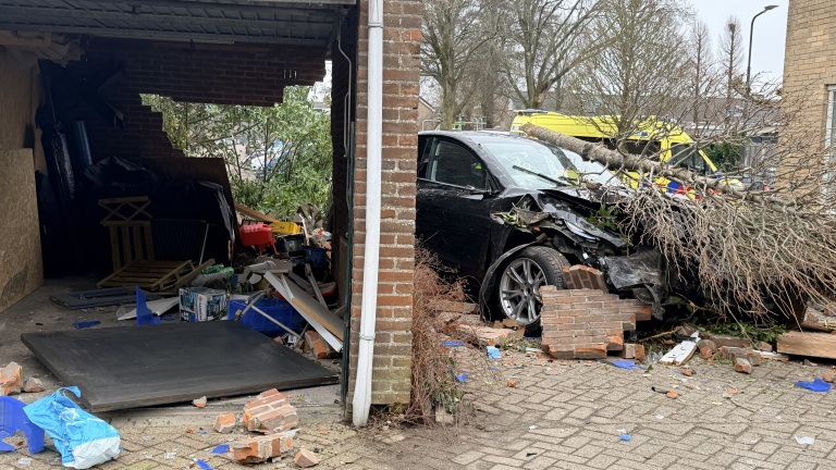 Een auto is gecrasht in een garage, omringd door puin en een omgevallen boom, met een gele reddingswagen op de achtergrond.