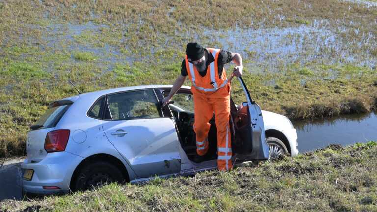 Politieauto en ambulance bij een ongeluk met een auto in een sloot langs een weg in een landelijk gebied.