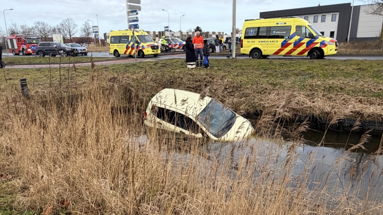 Auto half ondergedompeld in een sloot, met brandweer, ambulances en mensen op de achtergrond.