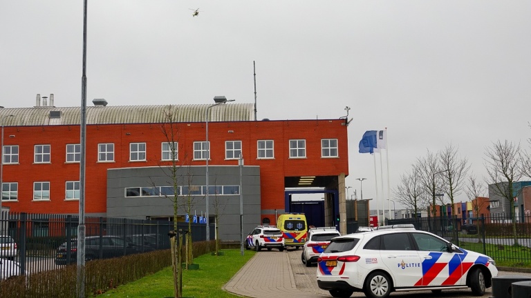 Politiewagens en een ambulance bij een rood bakstenen gebouw met vlaggen en een helikopter in de lucht.