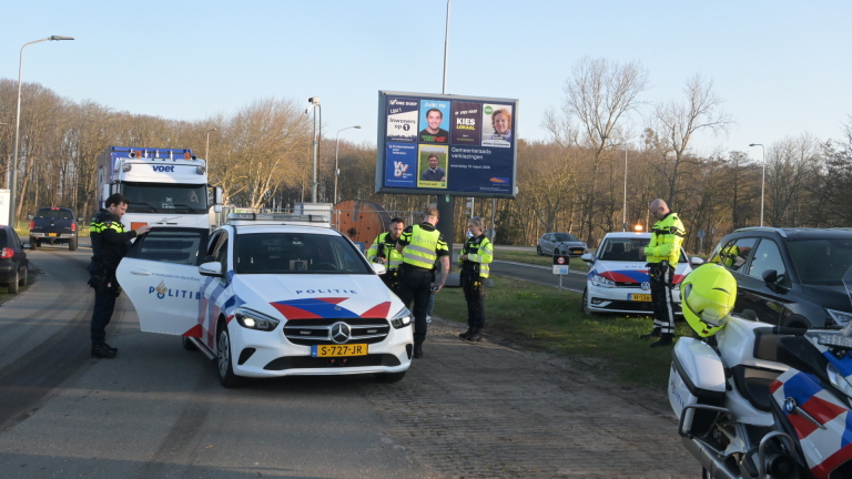Politieagenten staan naast meerdere politievoertuigen langs de weg, met een groot verkiezingsbord op de achtergrond.