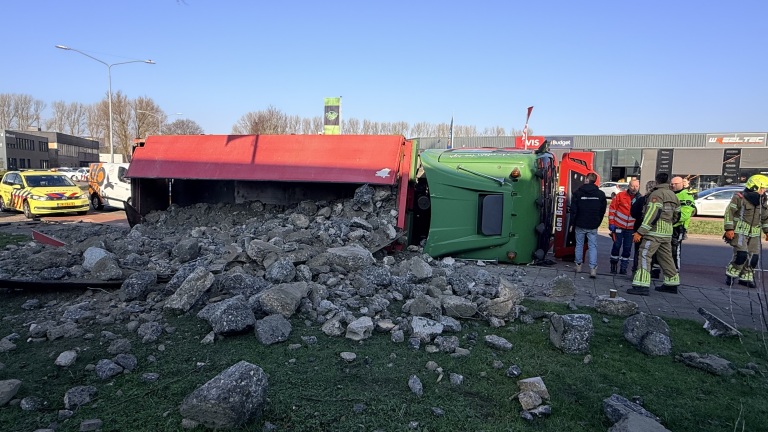 Een gekantelde vrachtwagen met verspreide stenen, omringd door hulpverleners in uniformen en voertuigen met zwaailichten.