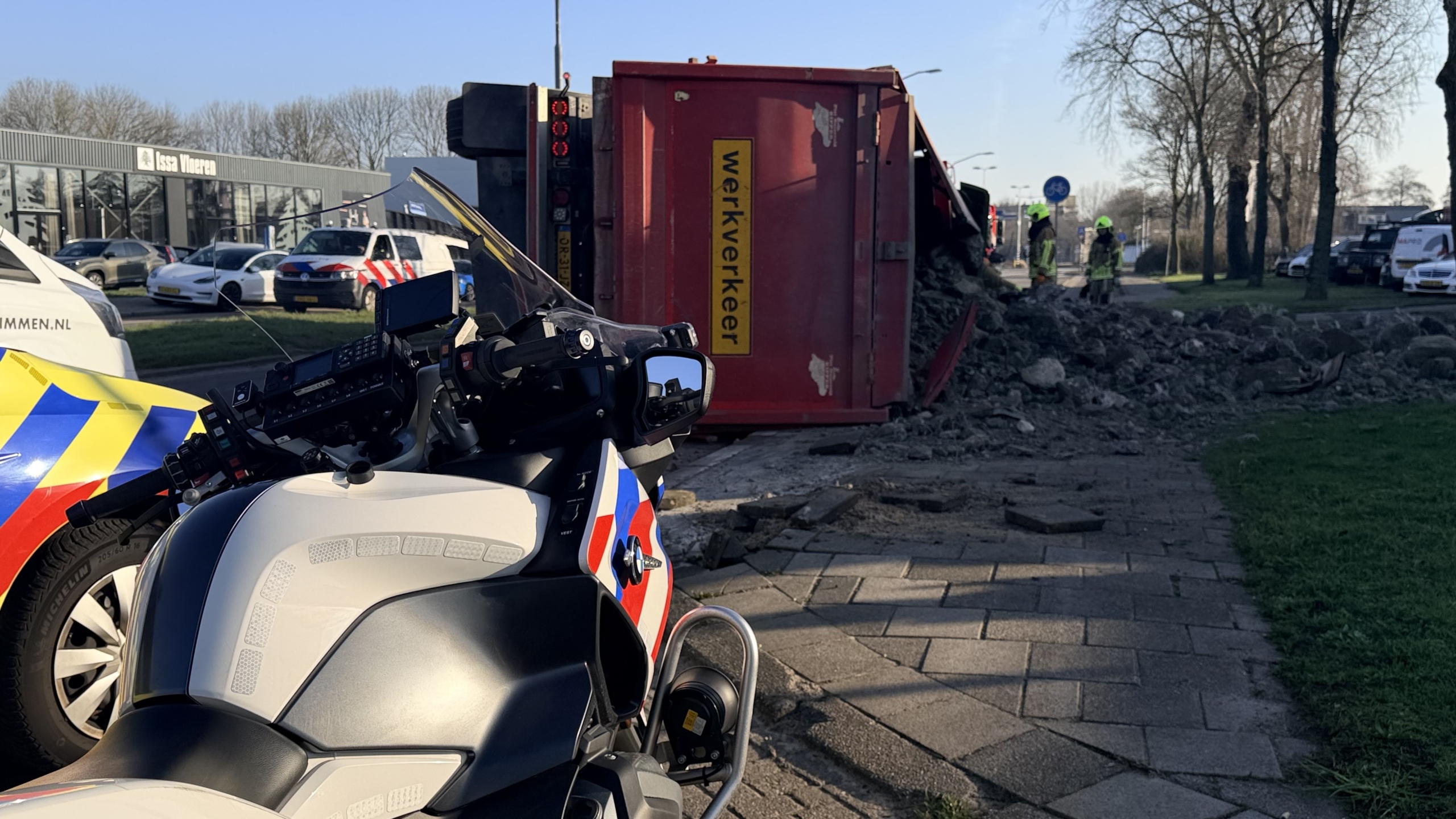 Een gekantelde vrachtwagen met verspreide stenen, omringd door hulpverleners in uniformen en voertuigen met zwaailichten.