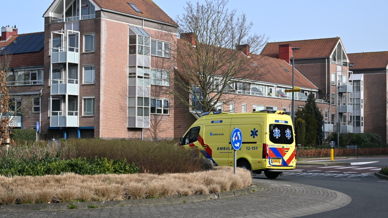 Gele ambulance en politie op motor bij een kruispunt naast flatgebouwen, met agenten en omstanders in gesprek.