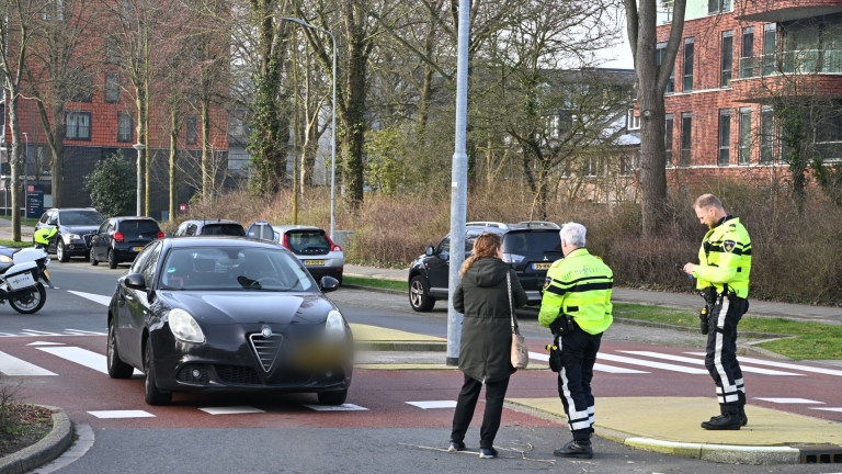 Gele ambulance en politie op motor bij een kruispunt naast flatgebouwen, met agenten en omstanders in gesprek.