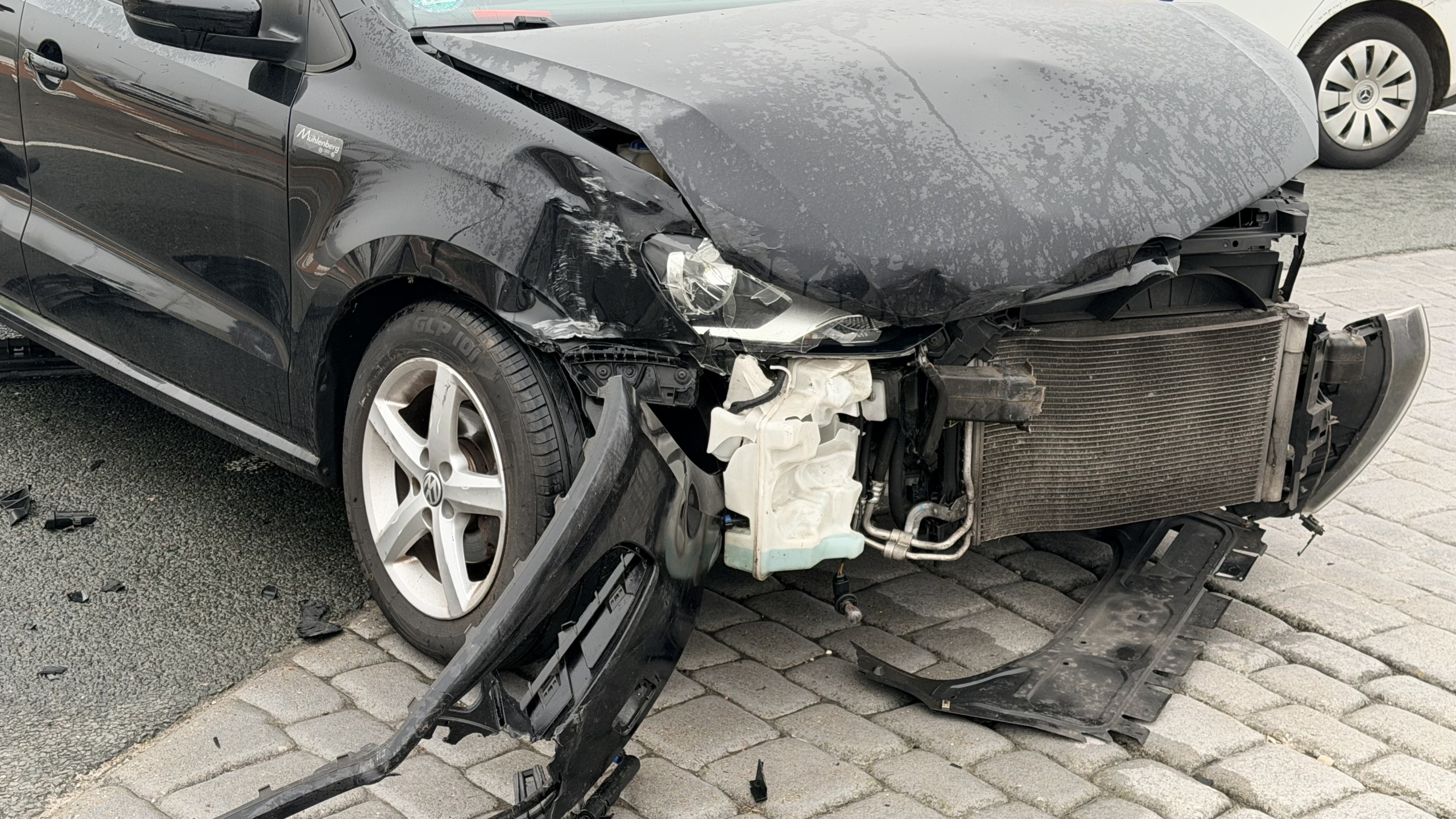 Auto met zware schade aan de voorzijde op een kruispunt, met politie en passerende voertuigen in de achtergrond.