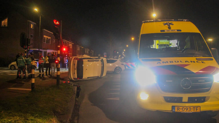 Een witte auto met schade naast een omgevallen voertuig op de weg in een nachtelijke setting, omstanders kijken toe.