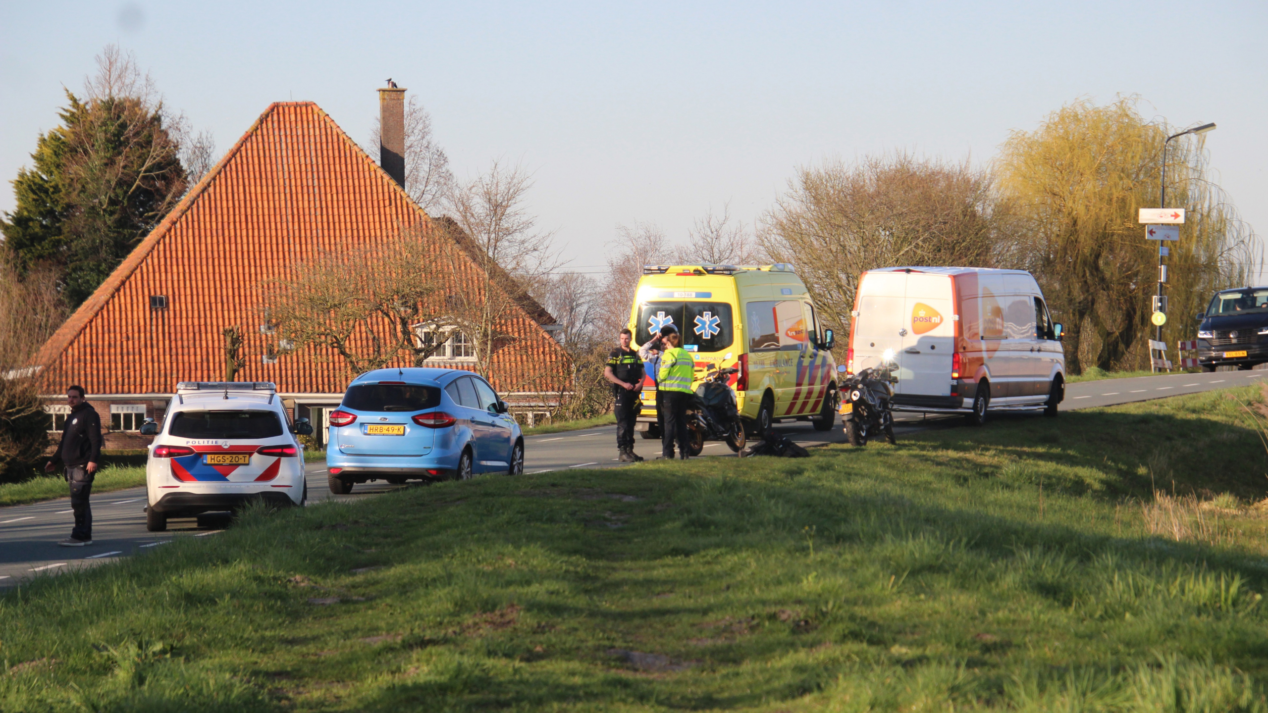 Ambulancepersoneel en politie staan bij een motor op een landelijke weg, terwijl een ambulance en auto's langs de weg stilstaan.