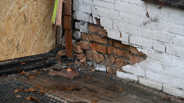 Vervallen gebouw met gebroken ramen en een dichtgetimmerde deur, omgeven door kapotte materialen op de grond.