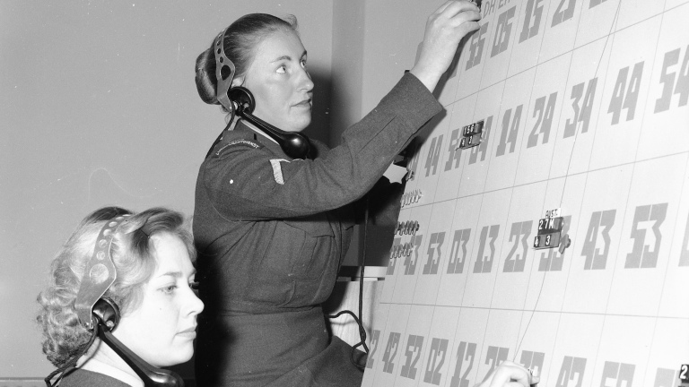 Twee vrouwen in uniform werken aan een groot bord met genummerde vakken, terwijl ze een telefoonkoptelefoon dragen.