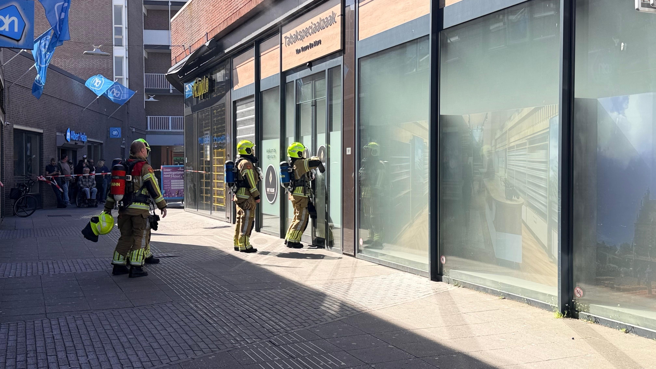 Brandweerlieden en politie bij een winkelstraat, met een politieagent op een motor en Albert Heijn vlaggen zichtbaar.