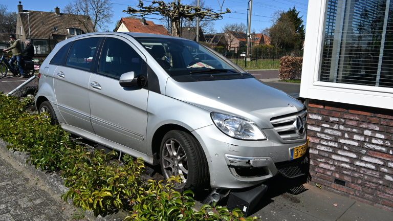 Een zilverkleurige auto is tegen een bakstenen muur en een tuinhek gebotst, met schade aan de bumper.