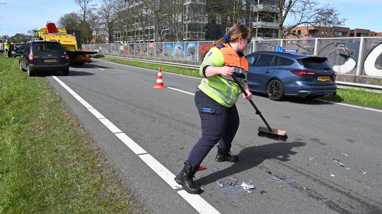 Een zwarte hatchback met een kapotte motorkap staat langs de weg, met politie en verkeer op de achtergrond.