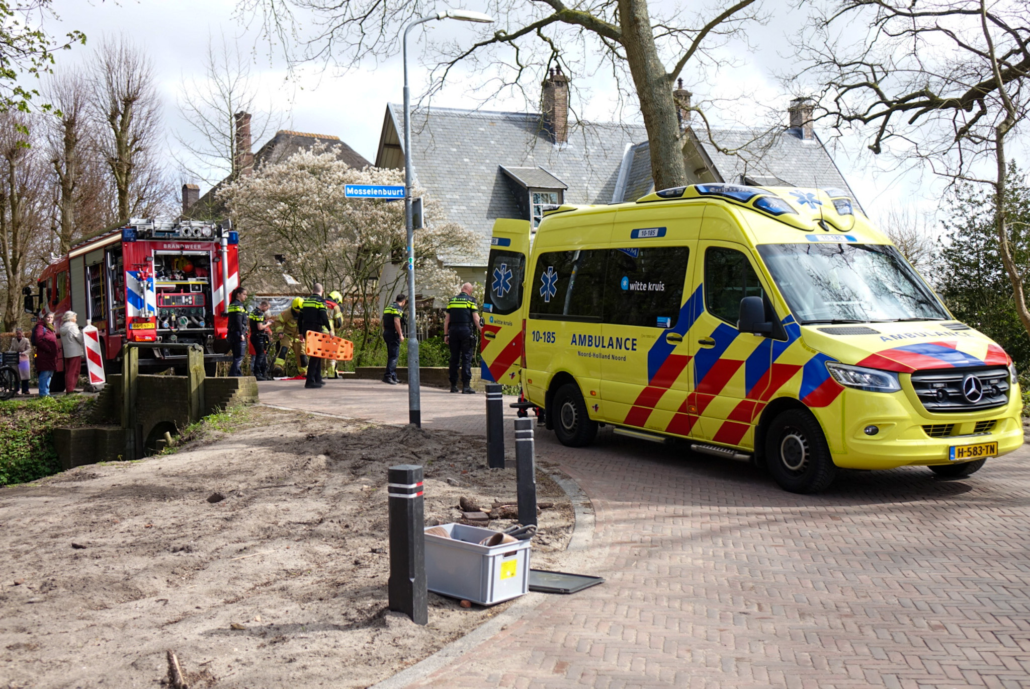 Gele ambulance en rode brandweerwagen bij wegafzetting in bosrijke omgeving; hulpdiensten aanwezig.