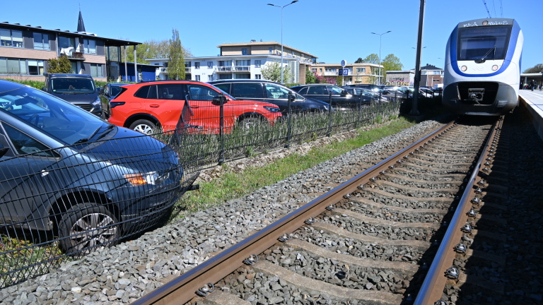 Auto tegen schuin hek met man in oranje vest die de deur opent. Politiewagen en omstanders op de achtergrond.