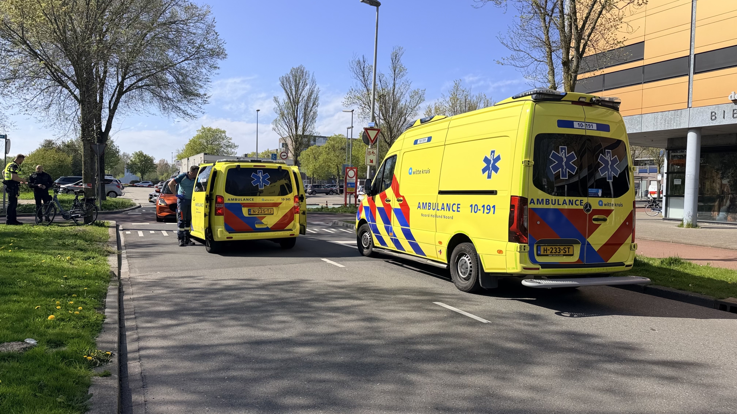 Een fiets is vastgebonden aan een boom naast een weg, naast een felgele auto en een oranje auto.