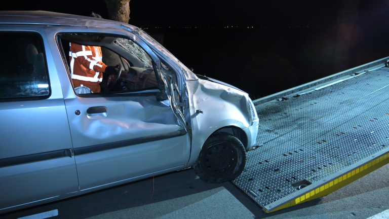Auto met schade aan een kant van de weg in het donker, met een persoon en politie in de buurt.