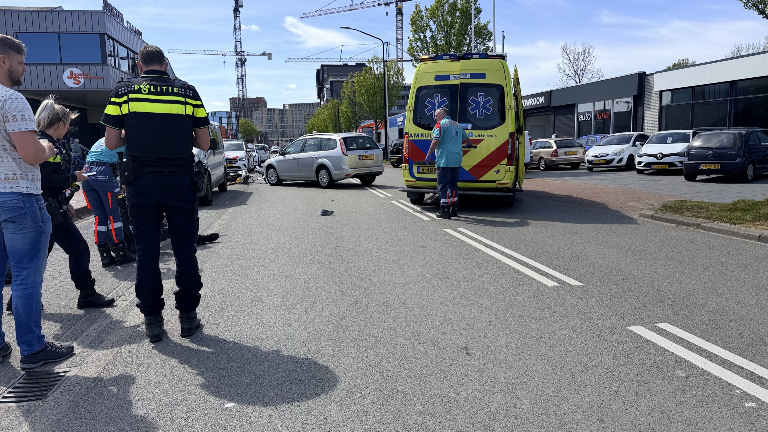 Politieagent onderzoekt een ongeval tussen een motorfiets en een grijze auto, nabij een showroom.