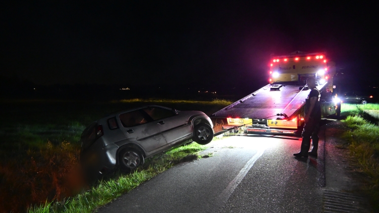 Een auto ligt gekanteld in een sloot in een grasrijke omgeving, met rook die uit de achterkant omhoog komt.