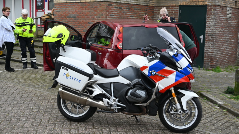 Agenten praten met een man naast een rode auto; een politiefiets met blauwe en rode strepen staat op de voorgrond.