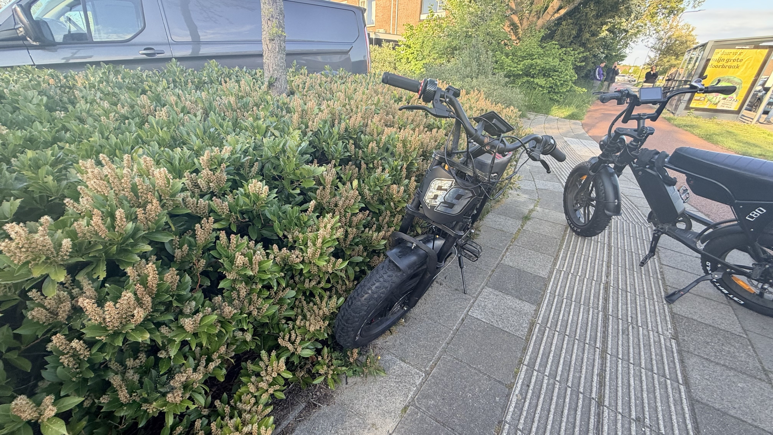 Groep mensen, waaronder politieagenten, verzameld rond een plek op straat met een bestelwagen en enkele elektrische fietsen op de achtergrond.