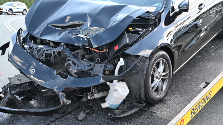 Twee auto's met schade na een botsing worden afgevoerd door een bergingsvoertuig; er zijn mensen in veiligheidskleding aanwezig.