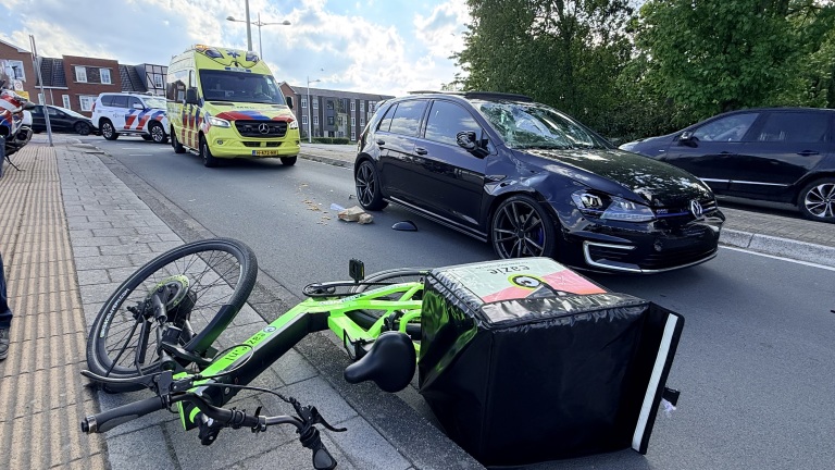 Omgevallen bezorgfiets naast een zwarte auto op de straat met een ambulance en politieauto op de achtergrond.