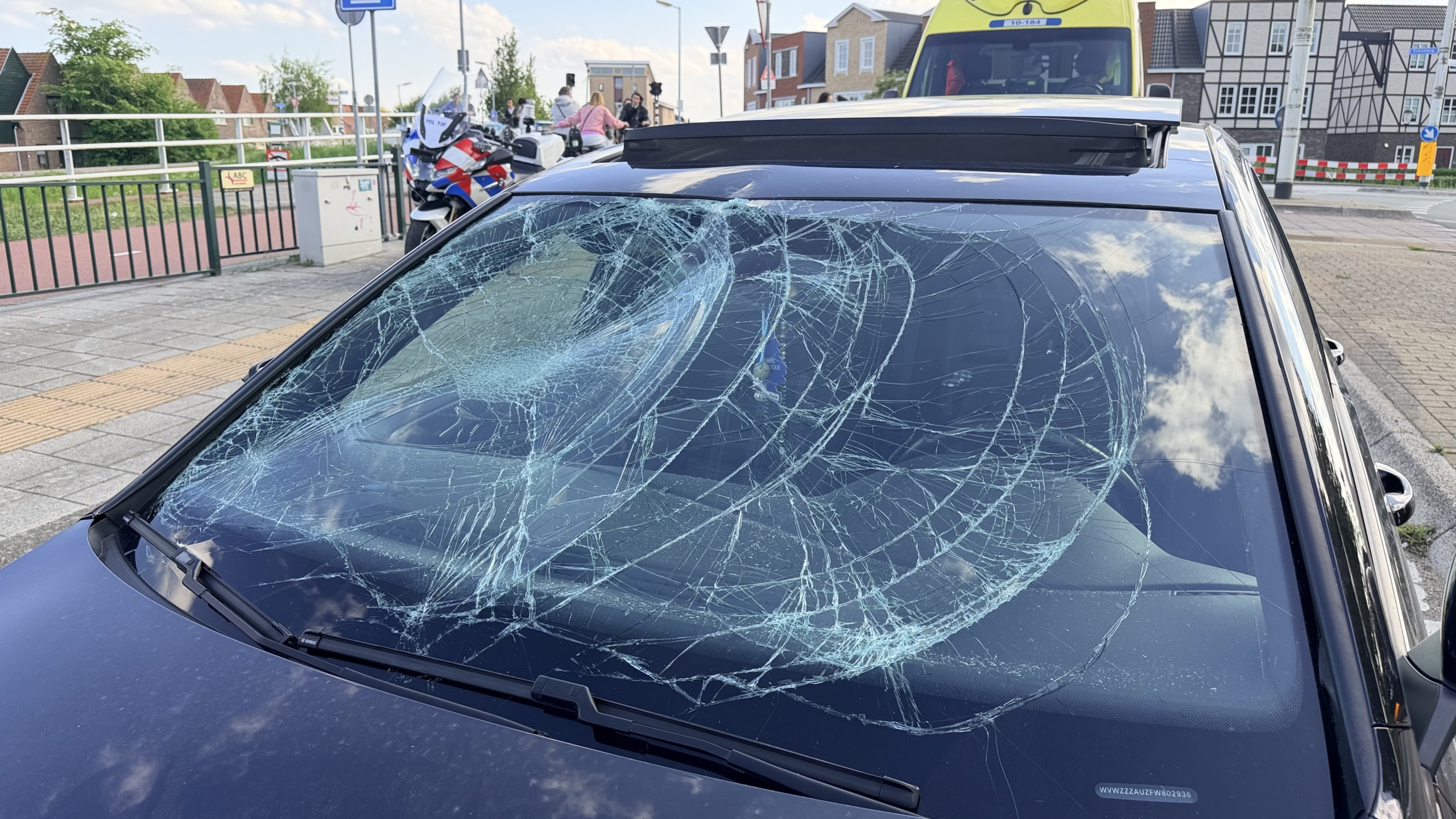 Omgevallen bezorgfiets naast een zwarte auto op de straat met een ambulance en politieauto op de achtergrond.