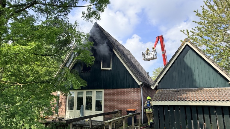 Brandweerlieden bestrijden brand in een huis met een gebroken raam en rook uit het dak, ondersteund door een hoogwerker.