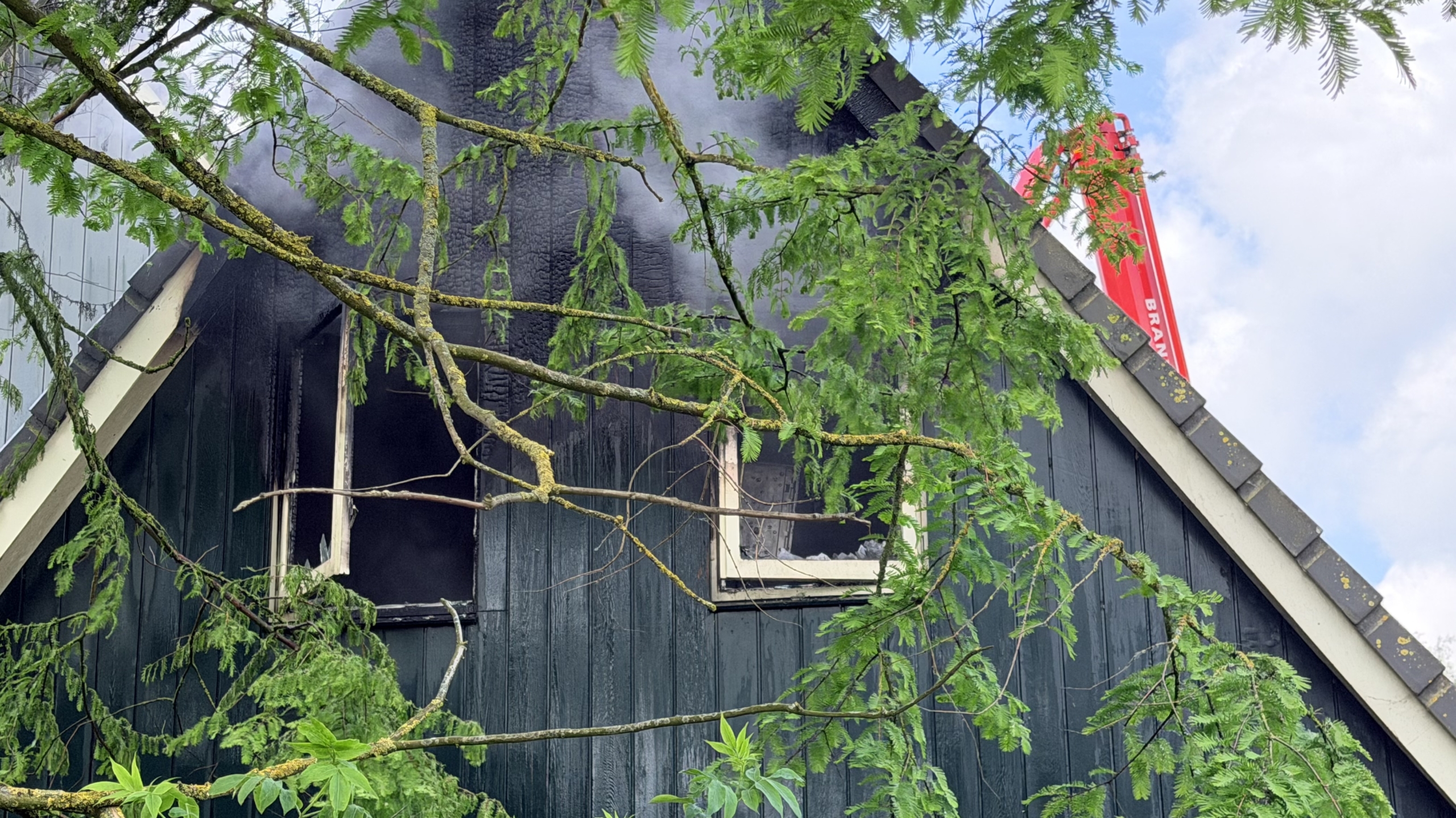 Brandweerlieden bestrijden brand in een huis met een gebroken raam en rook uit het dak, ondersteund door een hoogwerker.