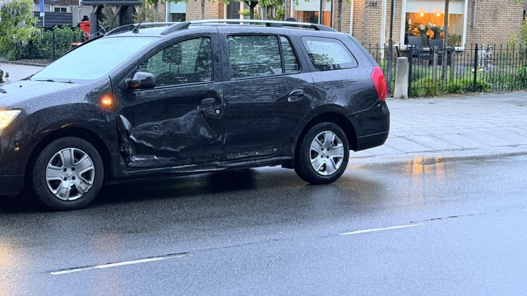 Zwarte auto met een grote deuk aan de zijkant, geparkeerd op een nat wegdek bij een rijtjeshuis.