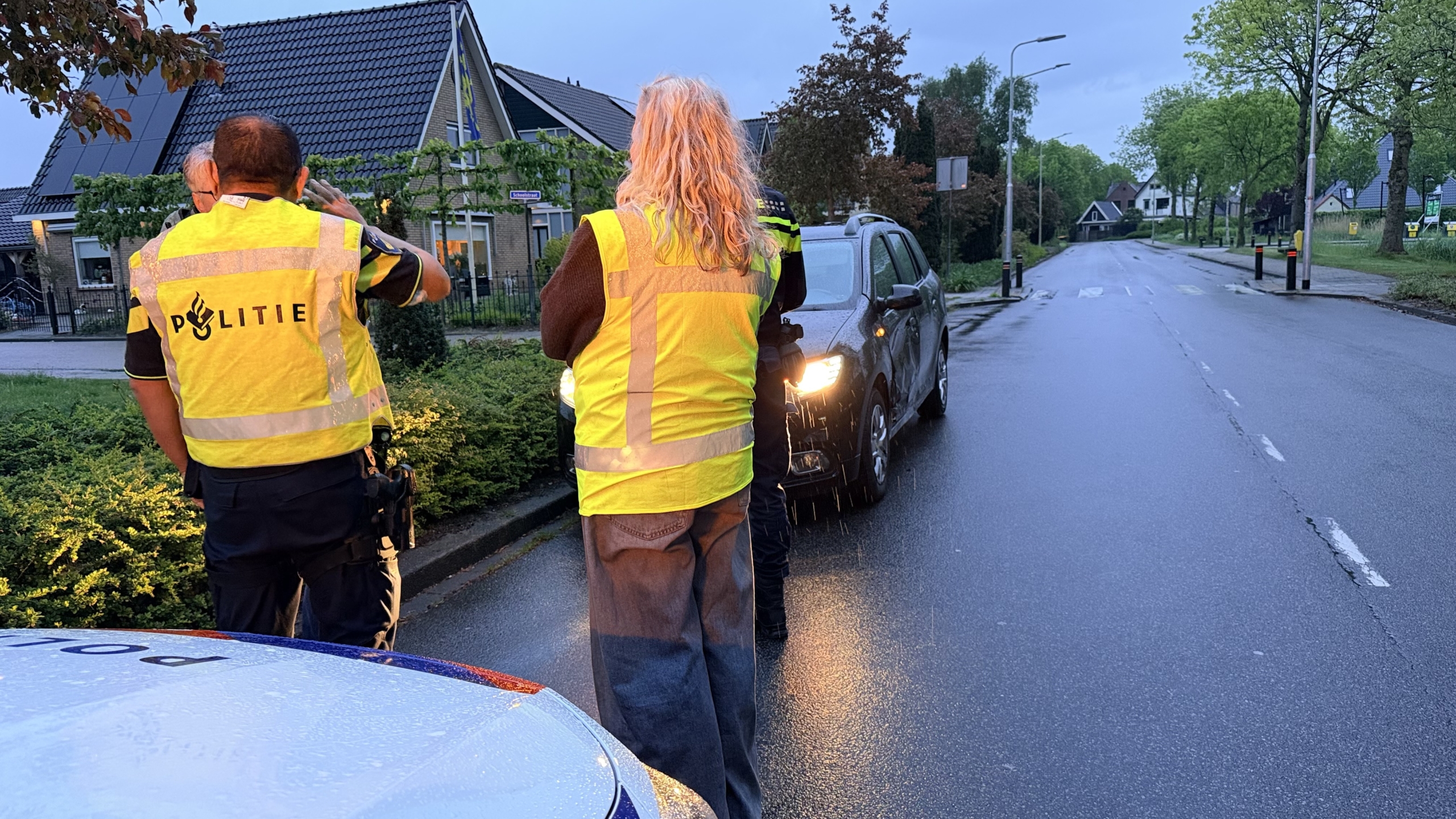 Zwarte auto met een grote deuk aan de zijkant, geparkeerd op een nat wegdek bij een rijtjeshuis.