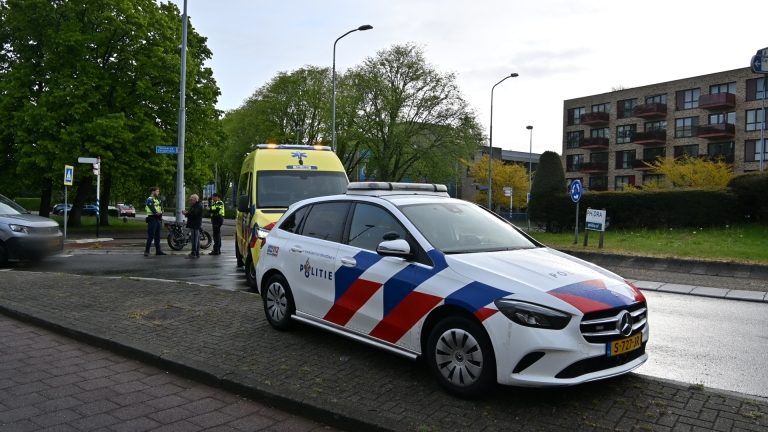 Politieauto en ambulance staan geparkeerd langs een weg in een stedelijke omgeving met huizen en een bus op de achtergrond.