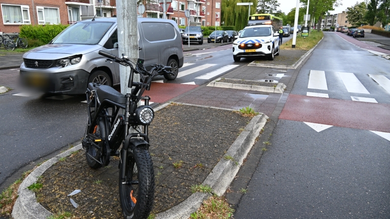 Politieauto en ambulance staan geparkeerd langs een weg in een stedelijke omgeving met huizen en een bus op de achtergrond.