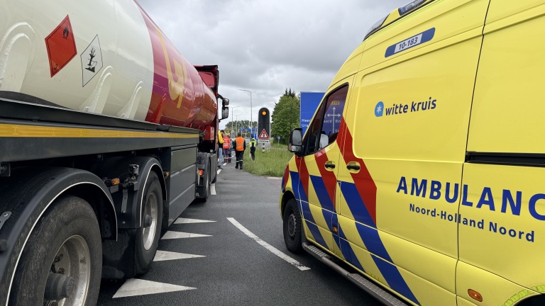 Een gele ambulance en een vrachtwagen zijn gestopt langs de weg, met mensen in veiligheidshesjes verderop.