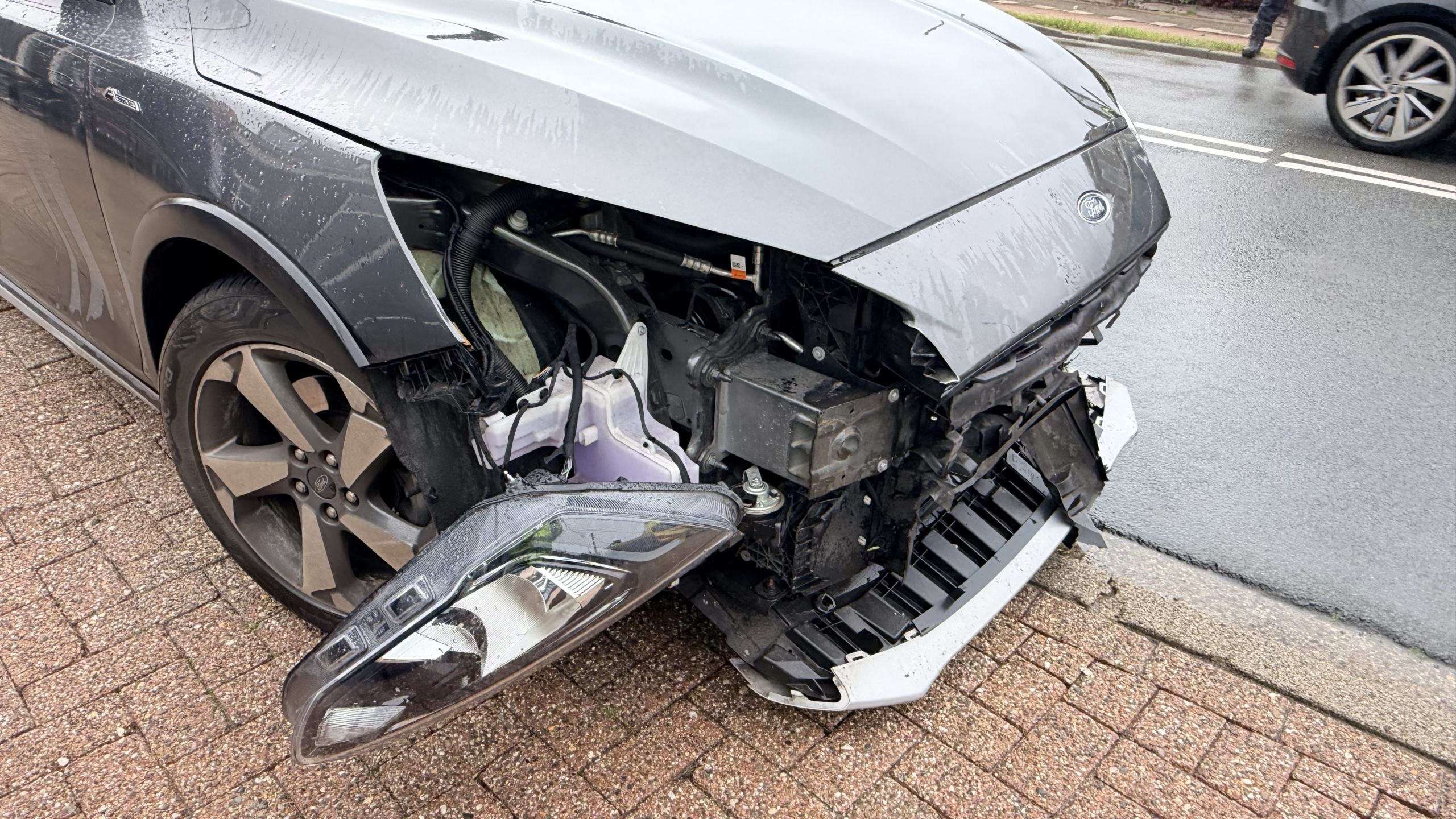 Een politieauto en een grijze auto met een kapot voorwiel staan op een natte weg voor een bergingsvoertuig.