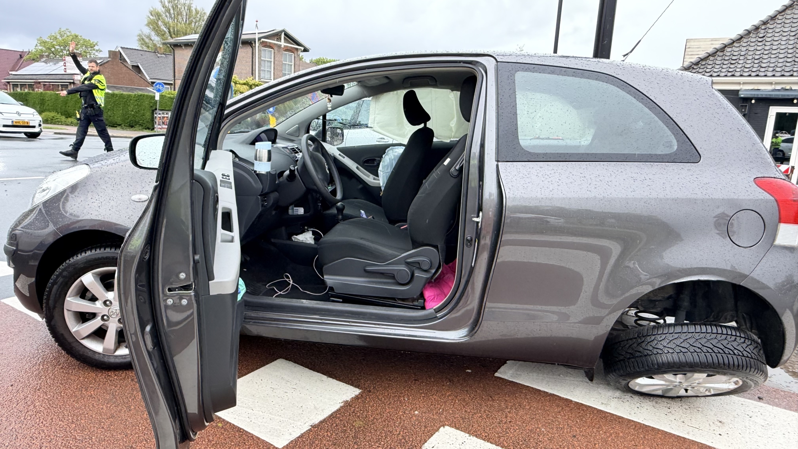 Een politieauto en een grijze auto met een kapot voorwiel staan op een natte weg voor een bergingsvoertuig.
